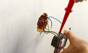 Instalator podłącza podwójny włącznik światła, używając próbnika napięcia. Widać przewody wychodzące ze ściany, gotowe do połączenia.