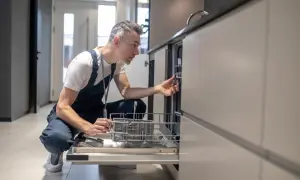 Mężczyzna w kombinezonie naprawczym klęczy i wyciąga koszyk ze zmywarki.