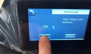 Palec dotyka ekranu wagi elektronicznej, inicjując kalibrację. Na wyświetlaczu widnieje komunikat "Połóż 1000g w celu kalibracji".