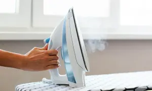 Dłoń trzyma parujące biało-niebieskie żelazko nad deską do prasowania. Jakie żelazko, takie gładkie ubrania!