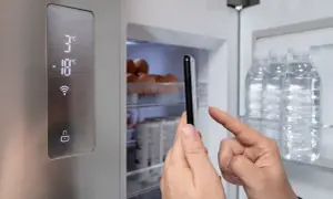 Lodówka Beko z cyfrowym wyświetlaczem i ikoną Wi-Fi. Ręka trzyma smartfon, kontrolując temperaturę. Czerwony wykrzyknik ostrzega o czymś.