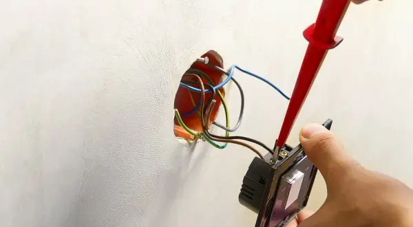 Instalator podłącza podwójny włącznik światła, używając próbnika napięcia. Widać przewody wychodzące ze ściany, gotowe do połączenia.