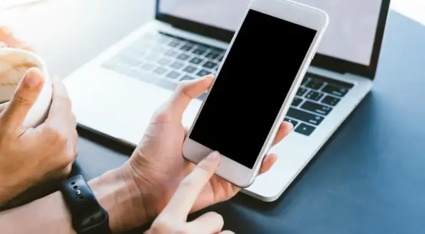 Dłoń trzyma biały smartfon, który nie chce się włączyć. Obok leży laptop, a w drugiej dłoni kubek z kawą.