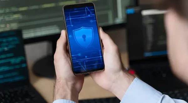 Ręce trzymają smartfon z ikoną tarczy i klucza, symbolizującą bezpieczeństwo. W tle widać monitory z kodem, sugerujące rozwiązanie problemu, jak zresetować zablokowany telefon.