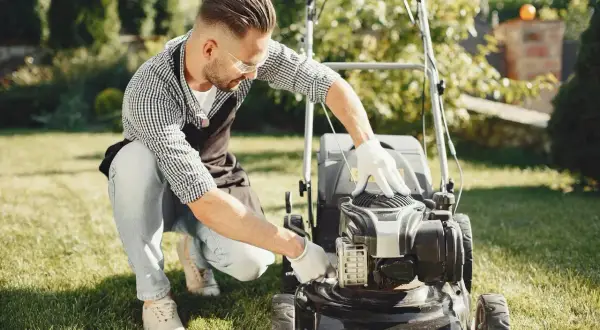 Mężczyzna w fartuchu i rękawiczkach próbuje uruchomić kosiarkę, która nie odpala.