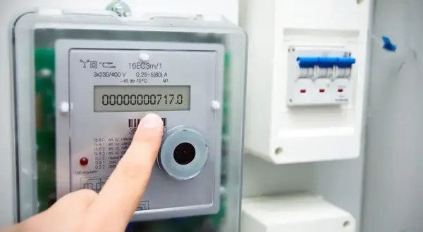Palec wskazuje licznik energii elektrycznej, na którym widnieje odczyt 717.0. Obok znajduje się rozdzielnica z bezpiecznikami. To ważne dla kontroli zużycia prądu i zarządzania rachunkami, a także dla zrozumienia kodów OBIS.