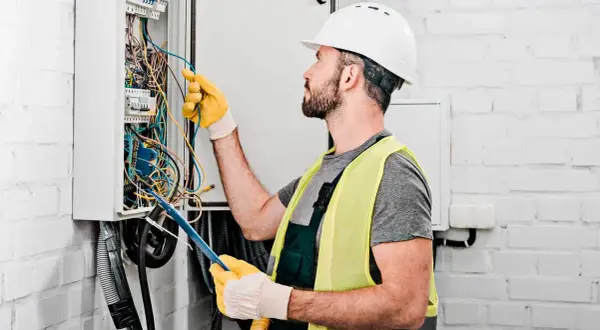 Elektryk w kasku i kamizelce odblaskowej pracuje przy skrzynce z przewodami. Zastanawia się, ile za punkt elektryczny.