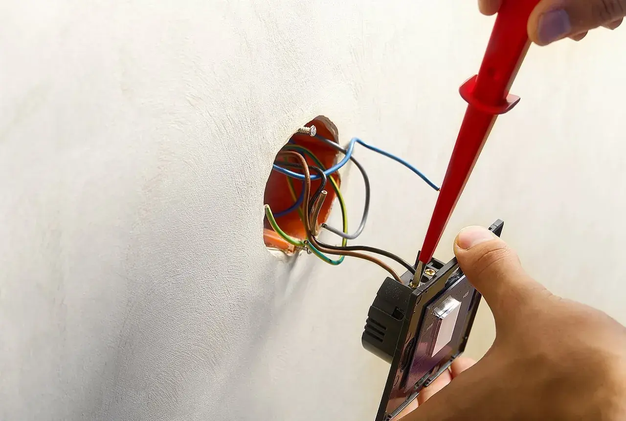 Instalator podłącza podwójny włącznik światła, używając próbnika napięcia. Widać przewody wychodzące ze ściany, gotowe do połączenia.