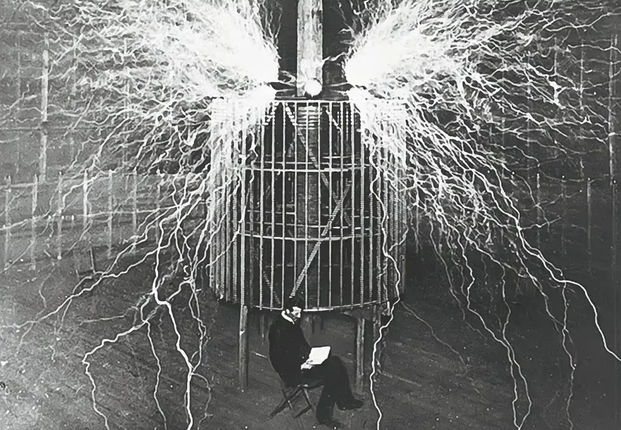 Nikola Tesla, siedzący w klatce, otoczony wyładowaniami elektrycznymi, pokazuje, kto wynalazł prąd.