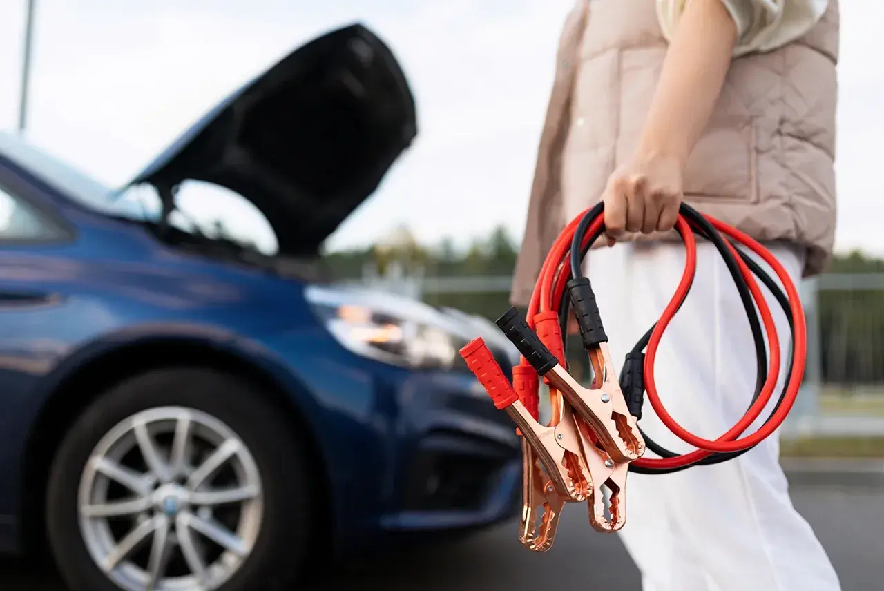 Osoba trzyma kable rozruchowe przy samochodzie z otwartą maską. Dowiedz się, jak podłączyć kable rozruchowe, by uruchomić auto.