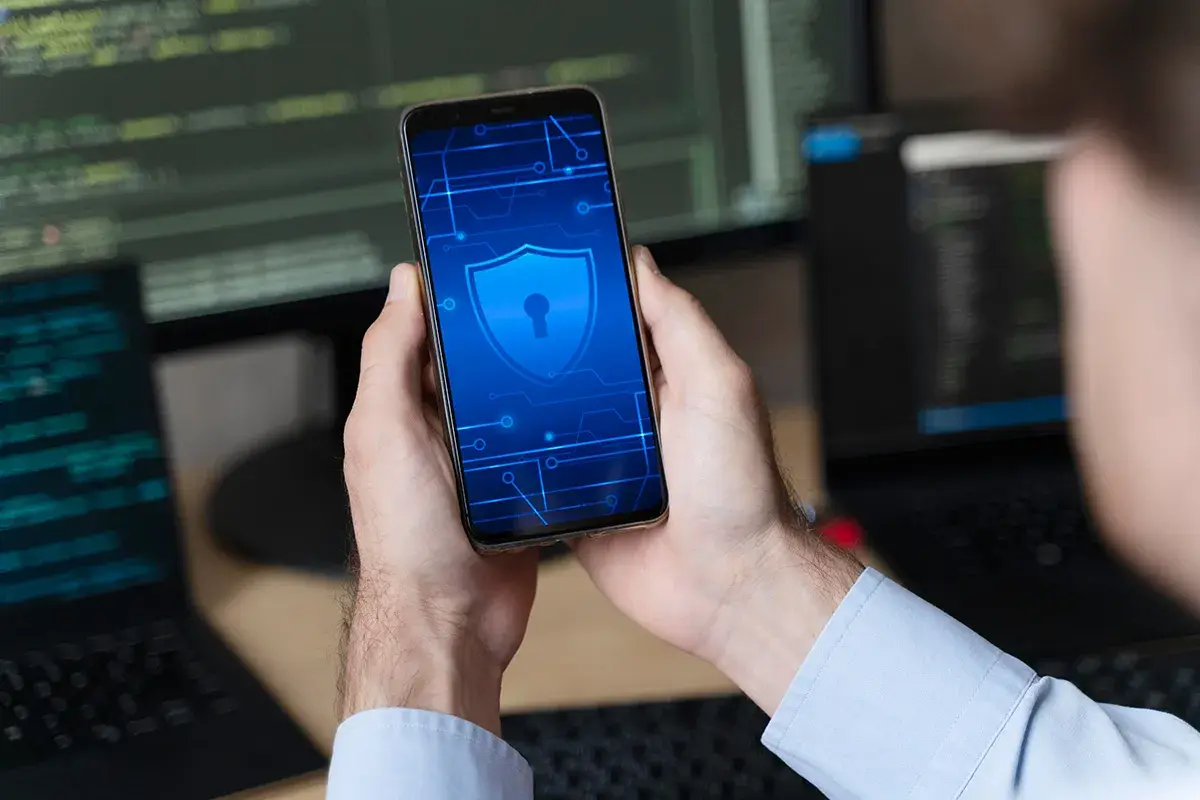 Ręce trzymają smartfon z ikoną tarczy i klucza, symbolizującą bezpieczeństwo. W tle widać monitory z kodem, sugerujące rozwiązanie problemu, jak zresetować zablokowany telefon.
