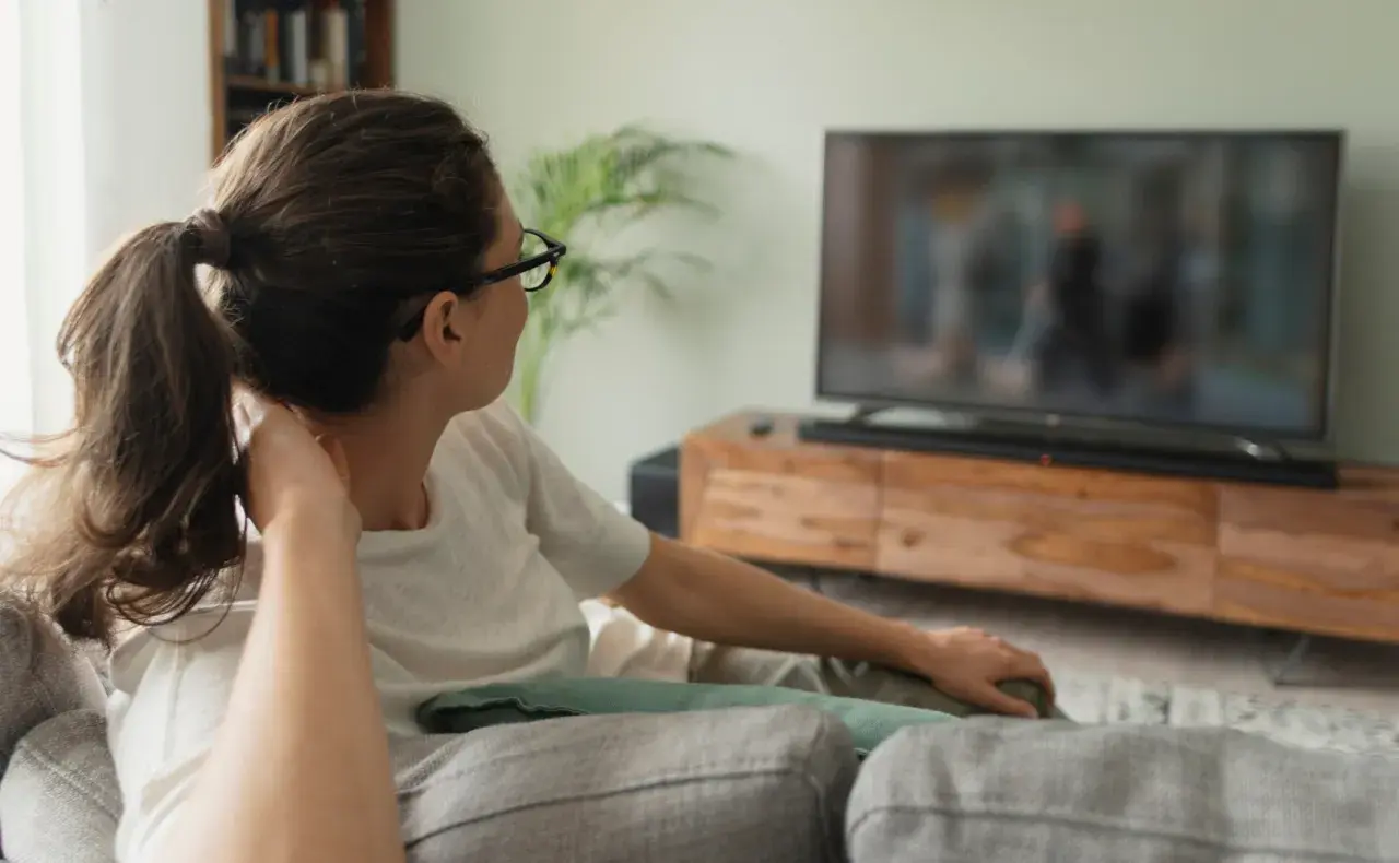 Kobieta w okularach relaksuje się na kanapie, oglądając telewizor. Idealna odległość od telewizora 65 cali zapewnia komfortowe wrażenia.