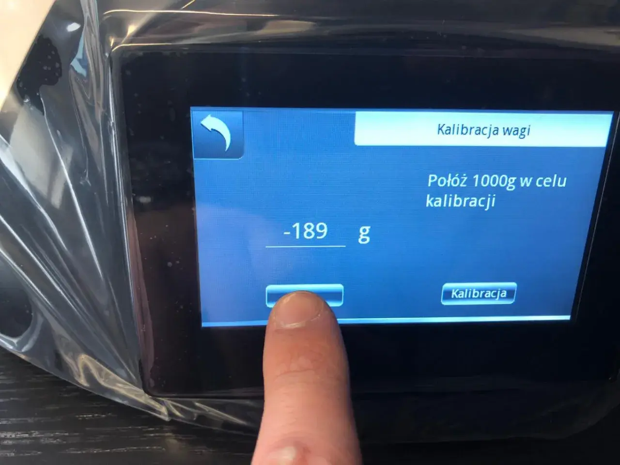 Palec dotyka ekranu wagi elektronicznej, inicjując kalibrację. Na wyświetlaczu widnieje komunikat "Połóż 1000g w celu kalibracji".