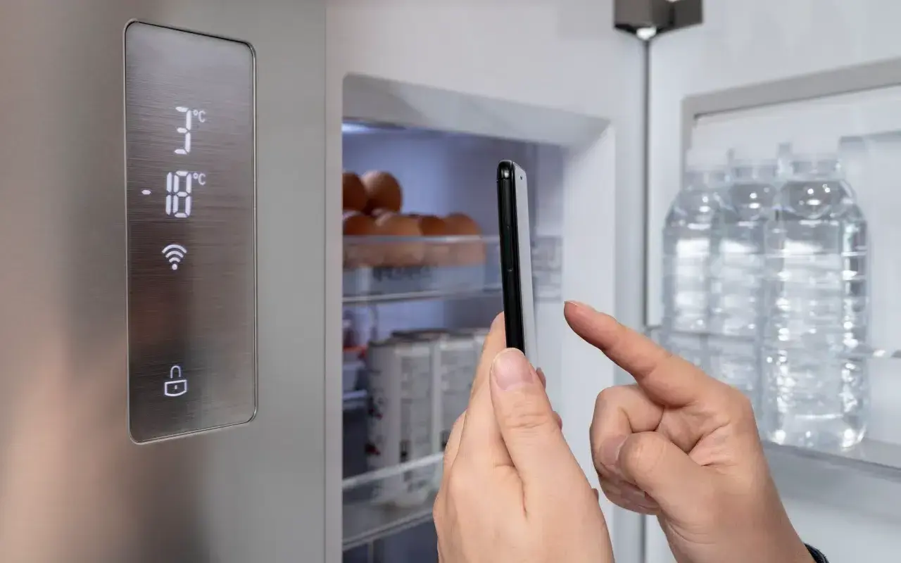 Lodówka Beko z cyfrowym wyświetlaczem i ikoną Wi-Fi. Ręka trzyma smartfon, kontrolując temperaturę. Czerwony wykrzyknik ostrzega o czymś.